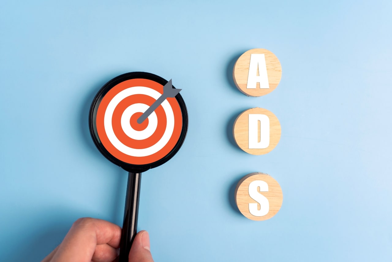 A magnifying glass focuses on a target with an arrow alongside wooden blocks spelling ADS.
