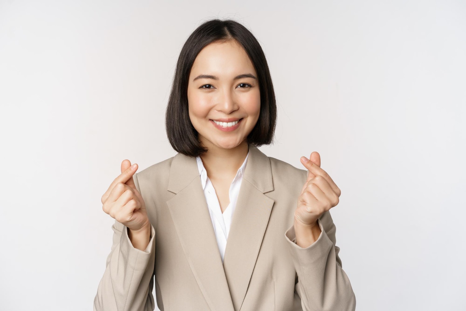Cheerful asian saleswoman smiling and showing finger hearts sign standing in suit over white background.
