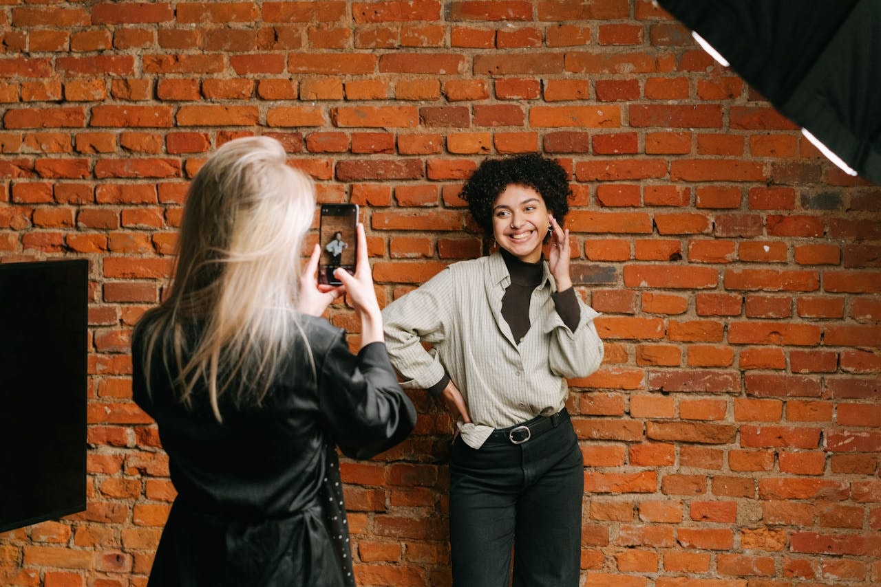 A woman taking a photo of a female model leaning against a brick wall