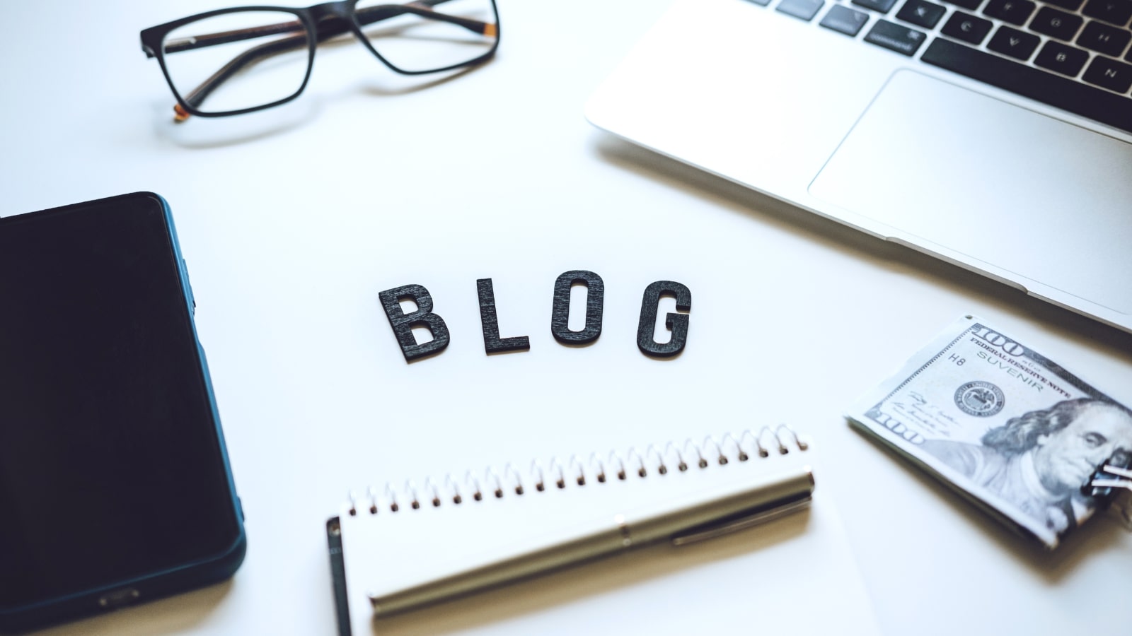 Blogging essentials on a desk like a phone, notebook, pen, and laptop.