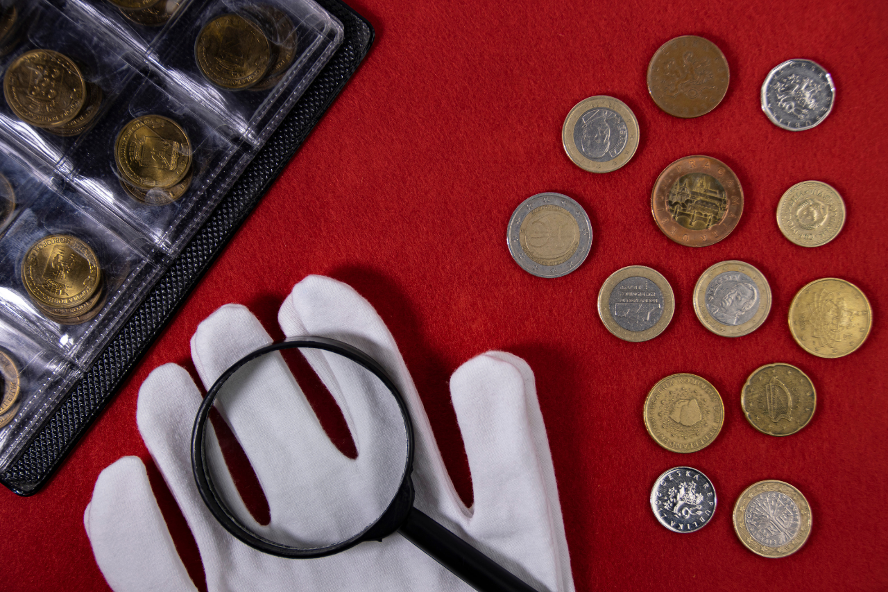 Collection of coins and collector white gloves and magnifying glass on red background.