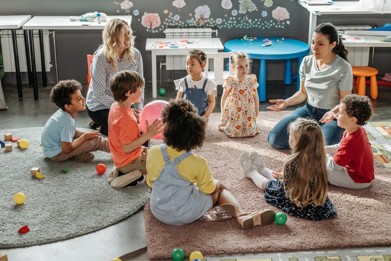 Two teachers with kids sitting on the floor, offering childcare services.
