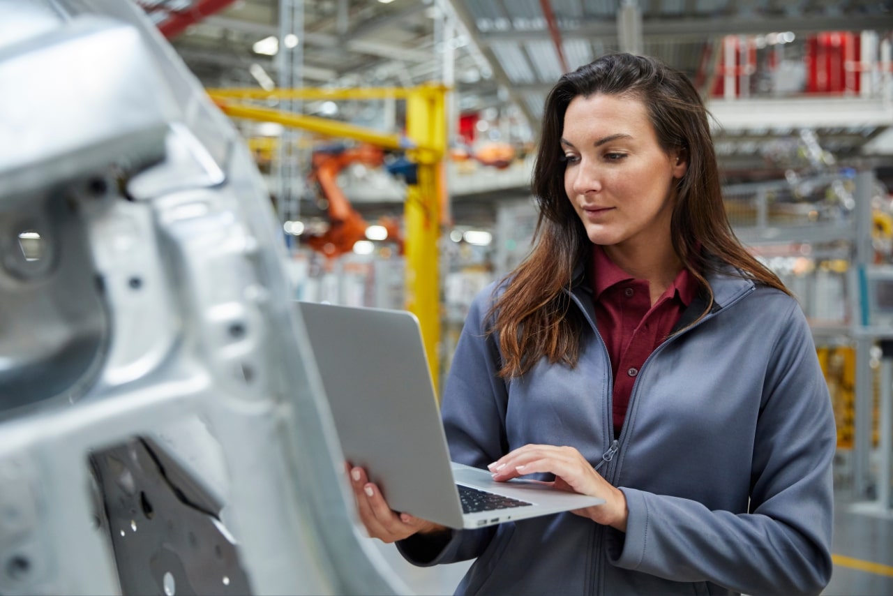 Woman engineer uses laptop in car factory to conduct predictive maintenance.