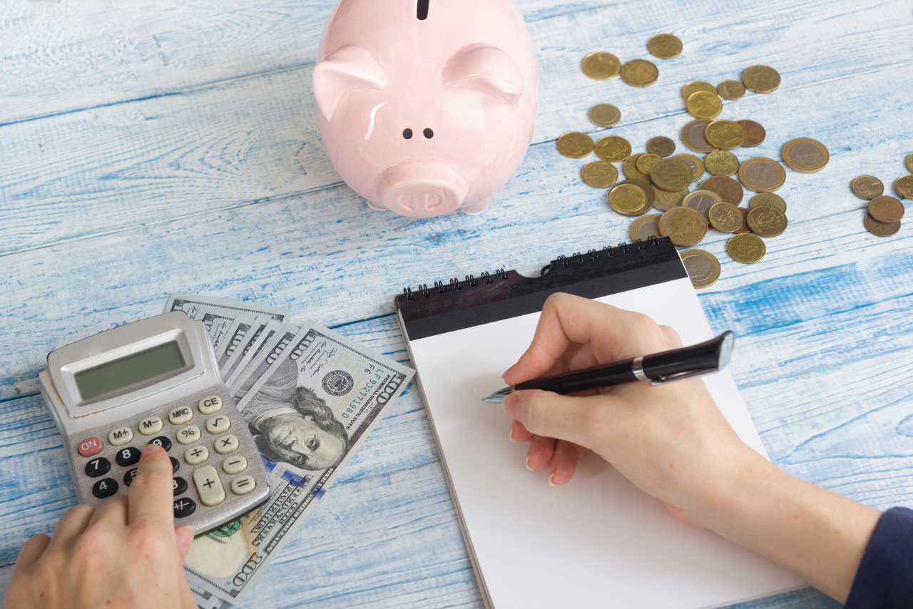 Woman writing on paper and computing amount saved with coins and cash on table.