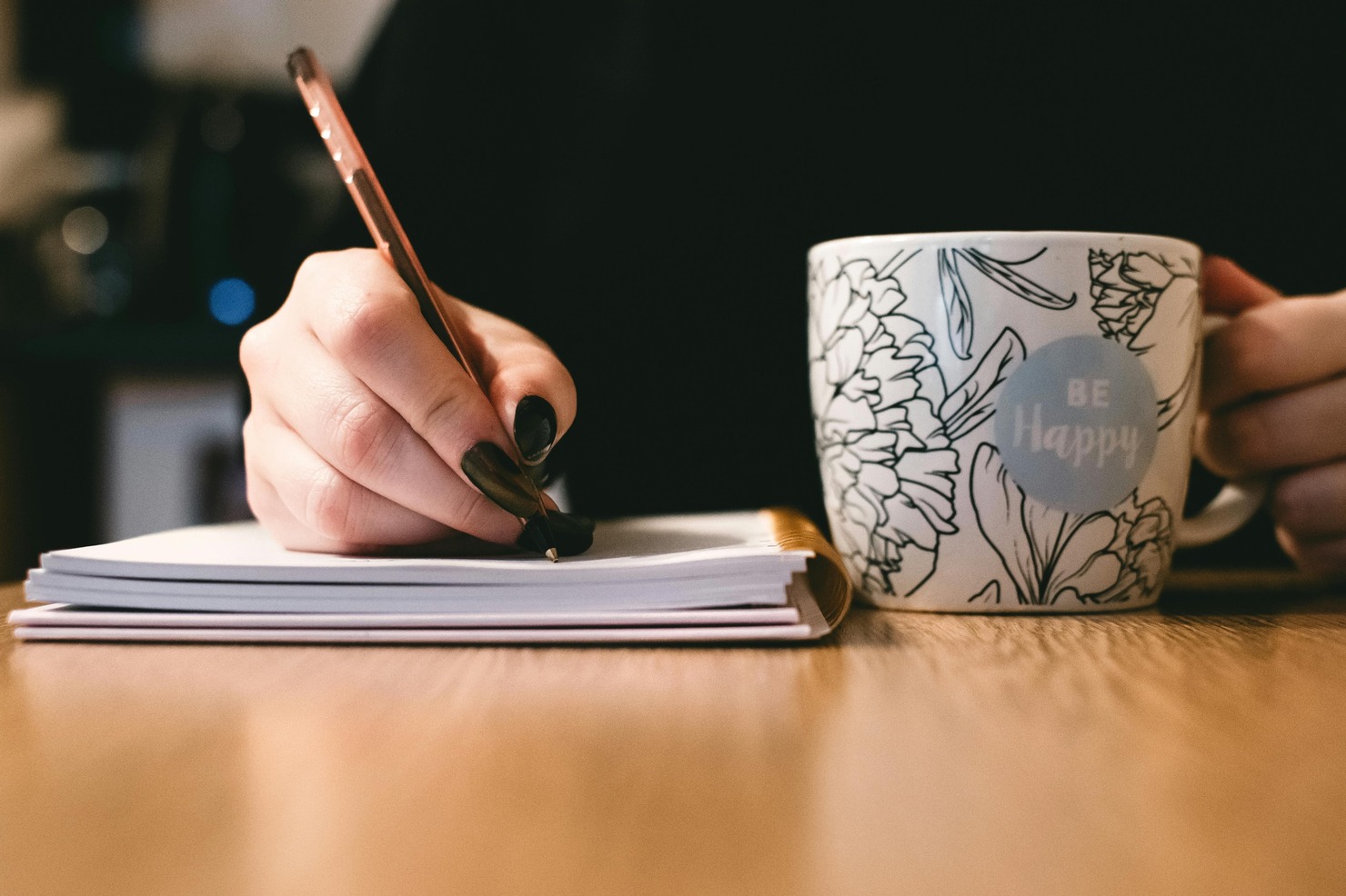A hand writes in a notebook while the other holds a cup.