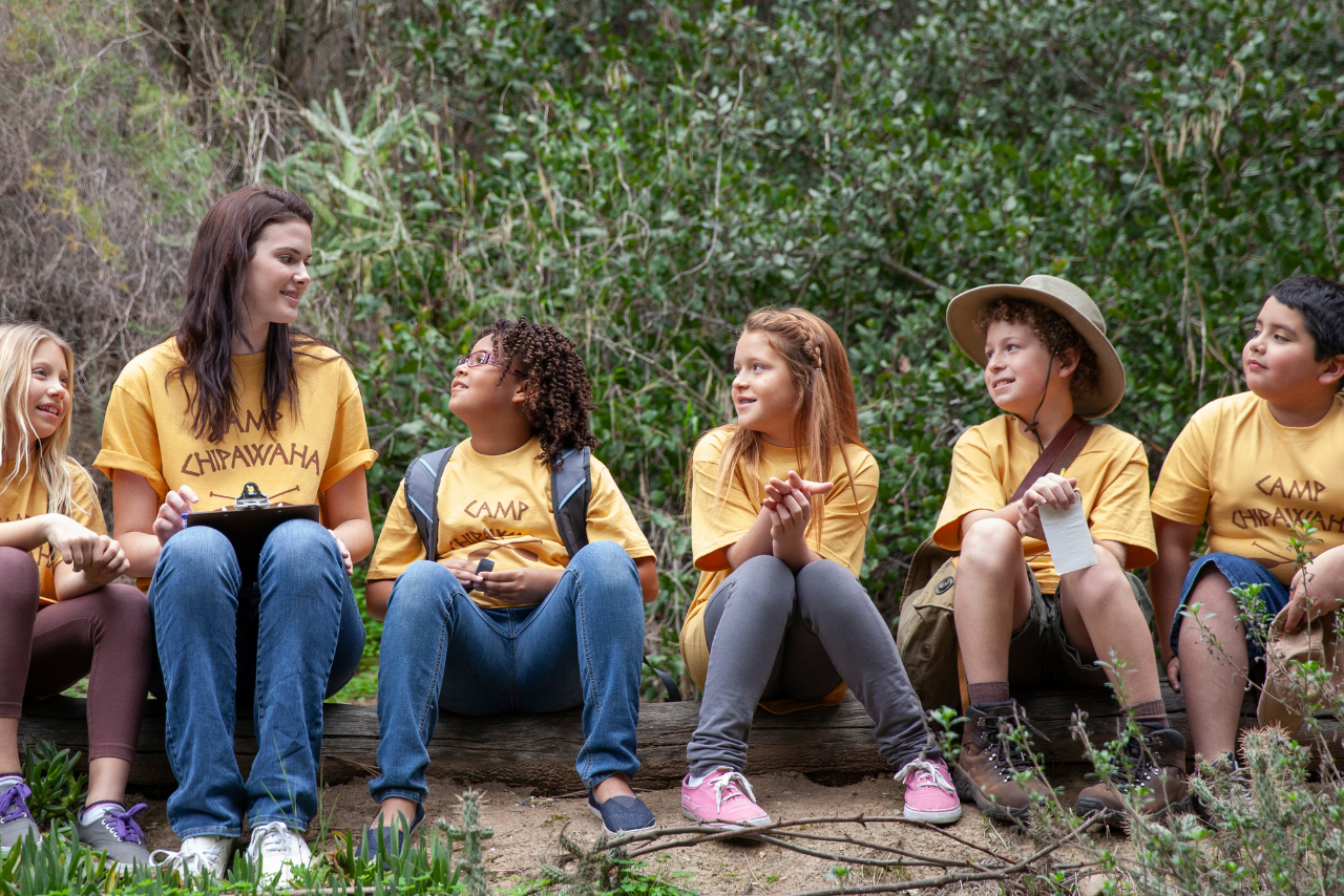 A summer camp counselor and kids sitting on log in woods.