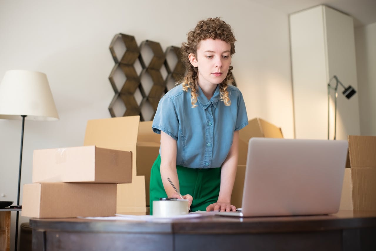 A woman surrounded by boxes looking at the computer and taking notes 