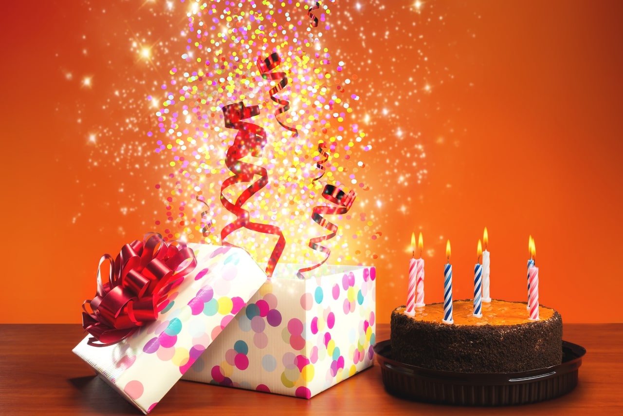 Birthday cake with candles and an opened gift with bursting glitters and red ribbons.