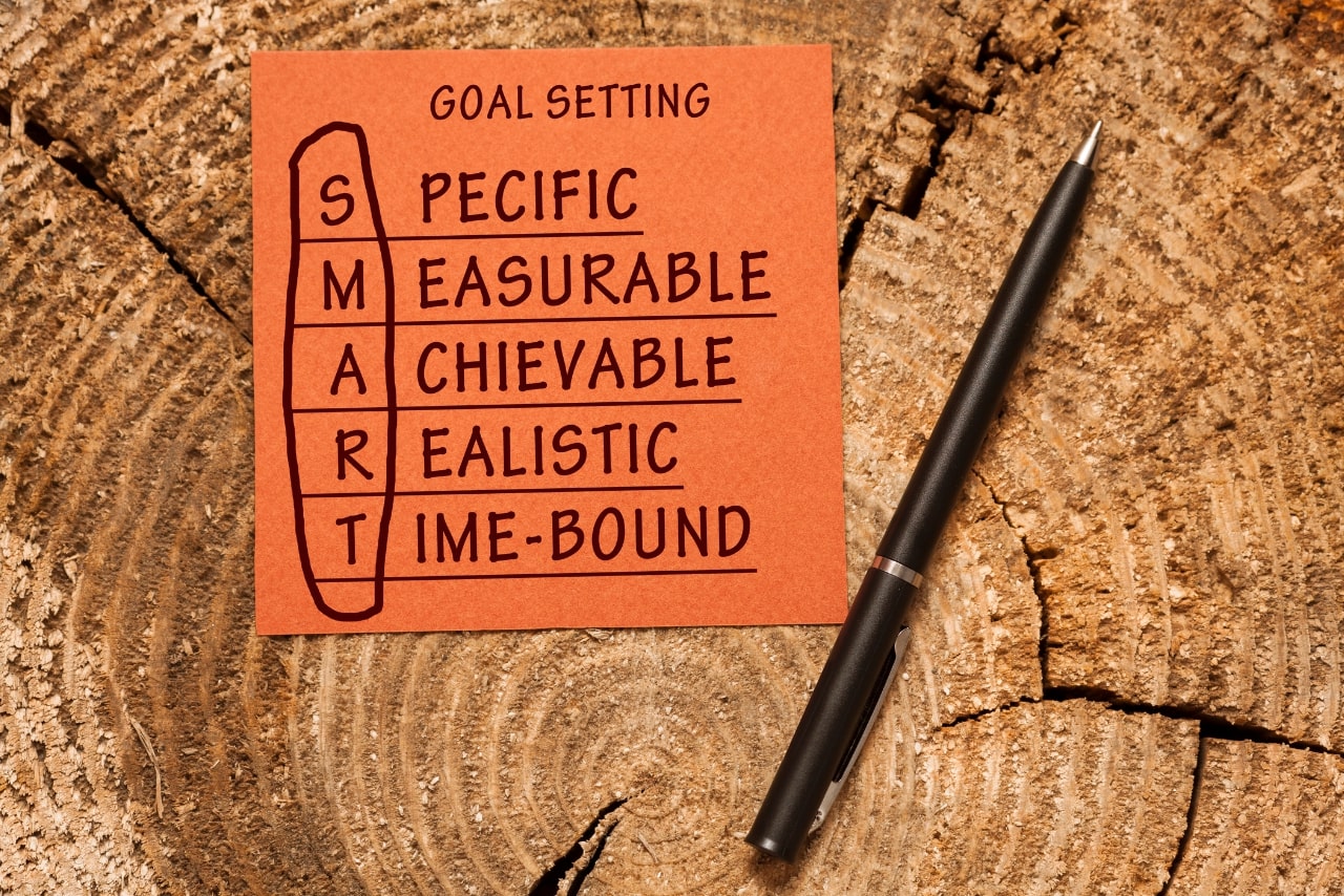 Pen and sticky note on a piece of wood with SMART acronyms (Specific, Measurable, Achievable, Realistic, Time-bound).