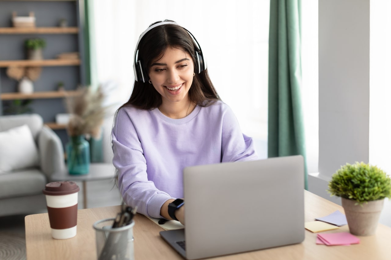 Smiling Woman Using Computer while Wearing Wireless Headphones.