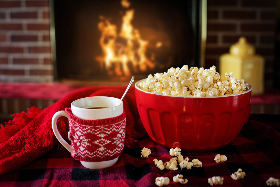 A coffee cup with a Christmas cozy sits beside popcorn, with a fireplace glowing in the background.