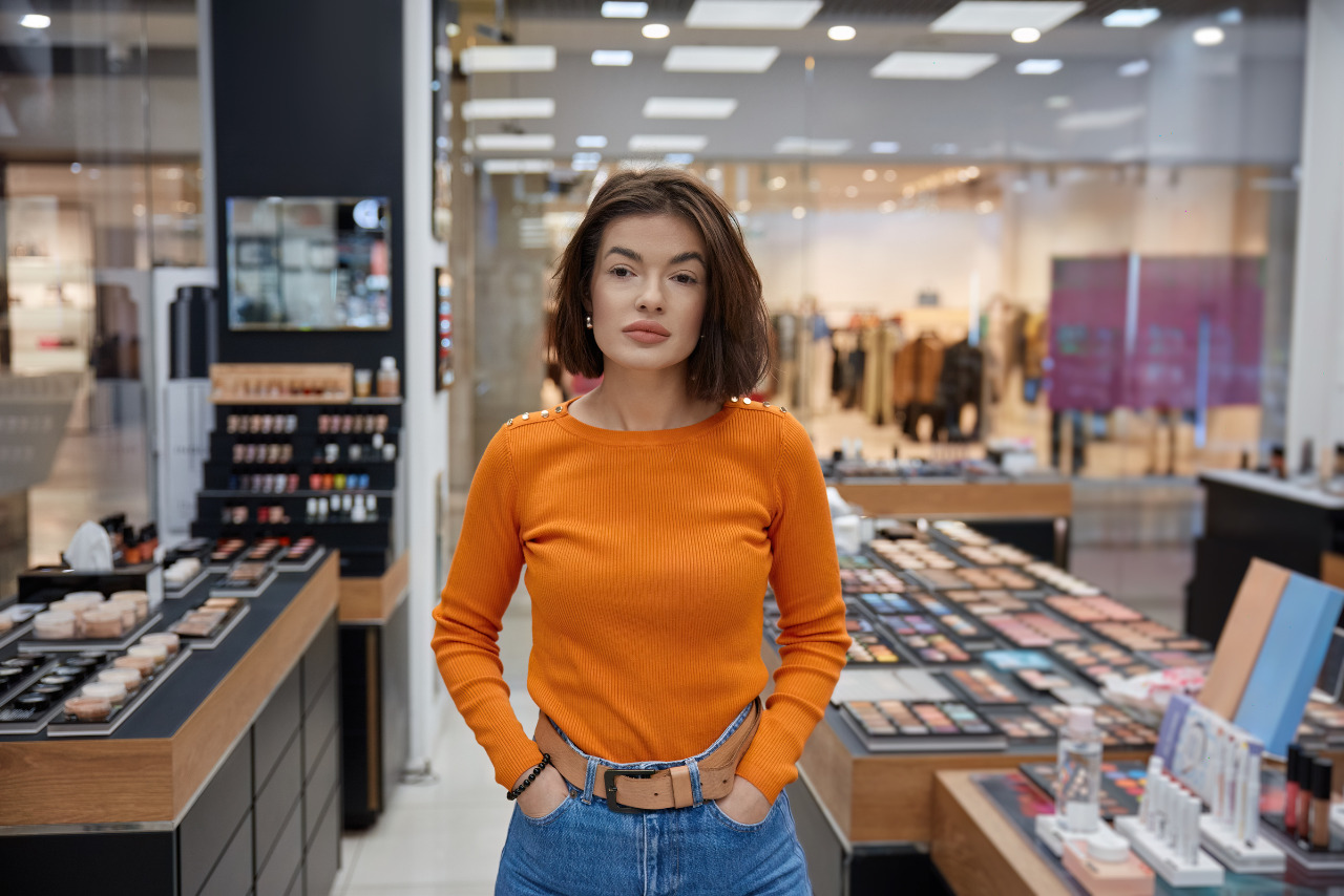 A woman in an orange sweater strolls through cosmetic display in a mall.