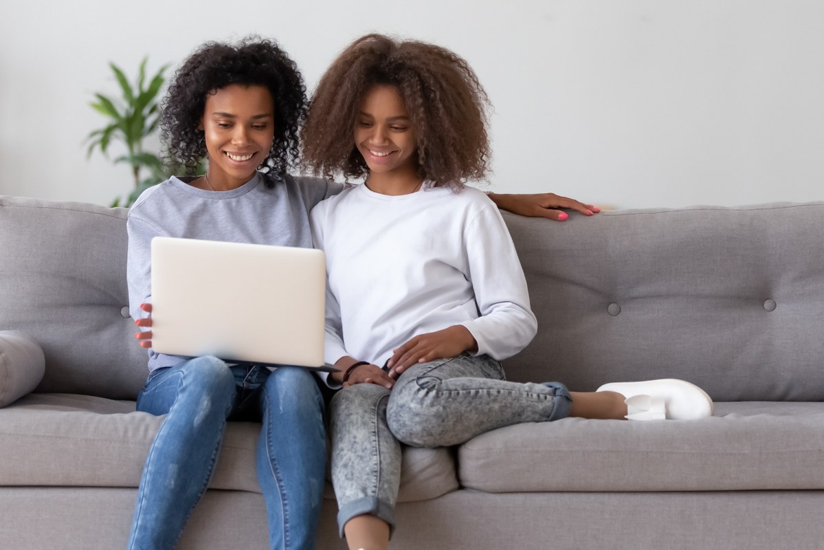 Happy African American mother and teen daughter using laptop on sofa.