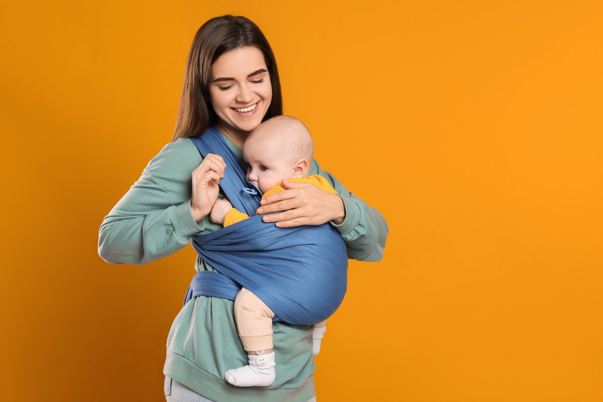 Mother holding her child in sling (baby carrier) on orange background.