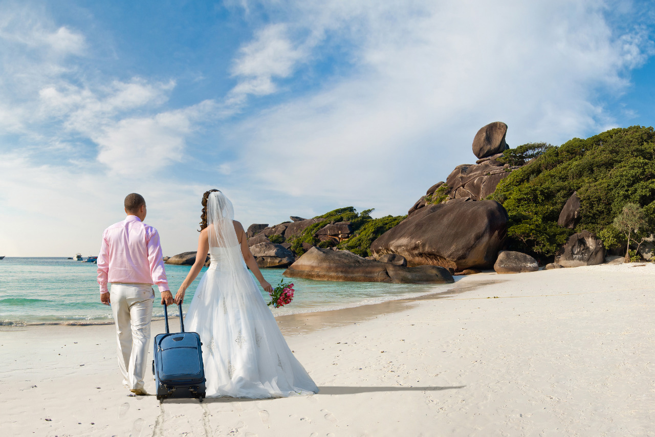 Newlyweds walking away on a sunny beach, carrying luggage.