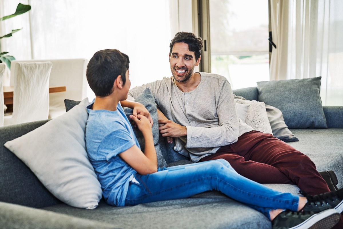 Son listens attentively and respectfully while father talks to him on sofa at home.