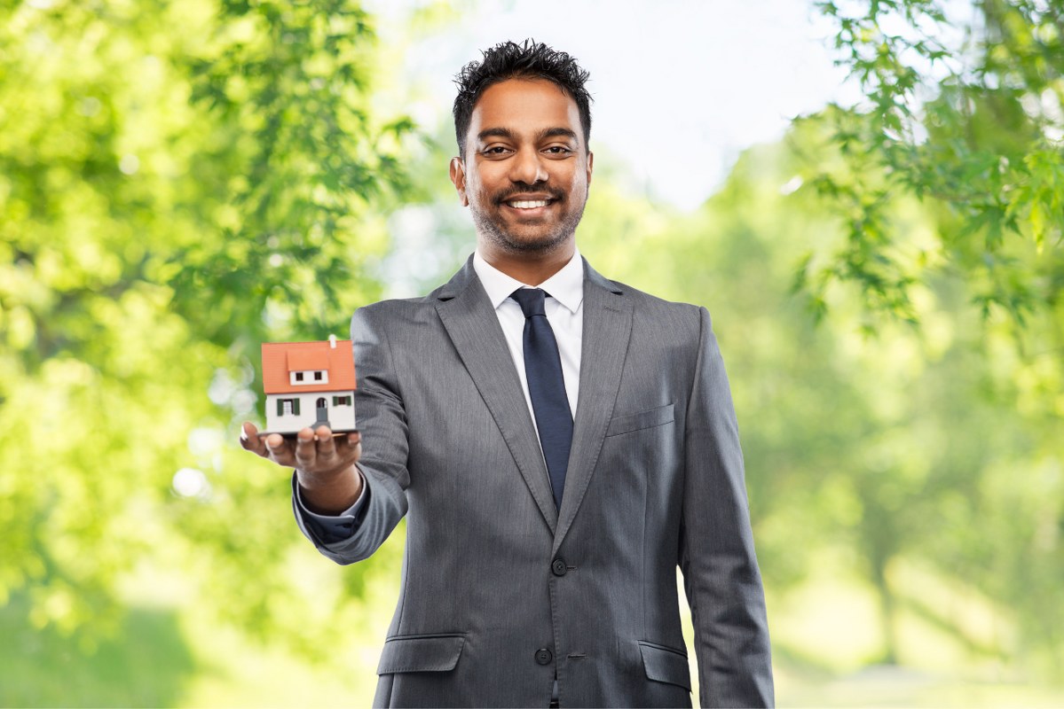 Indian businessman stands while holding miniature house.