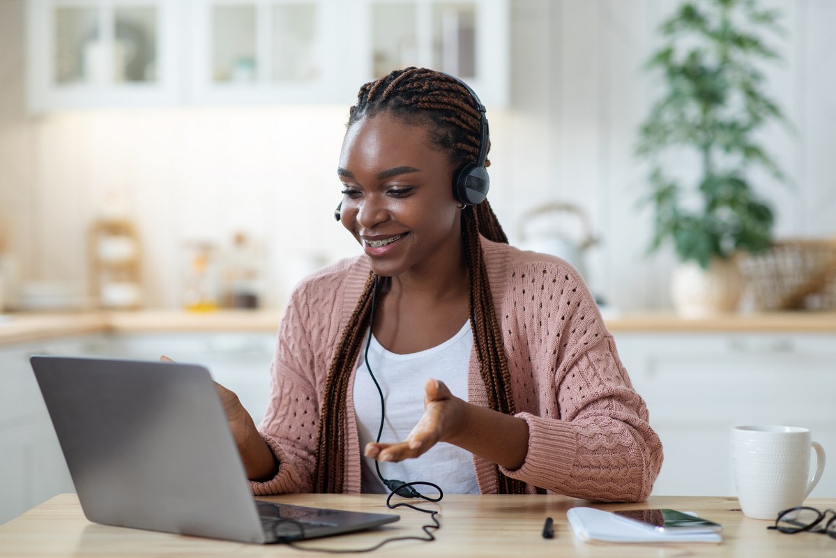 Smiling black female tutor in headset using laptop in kitchen.