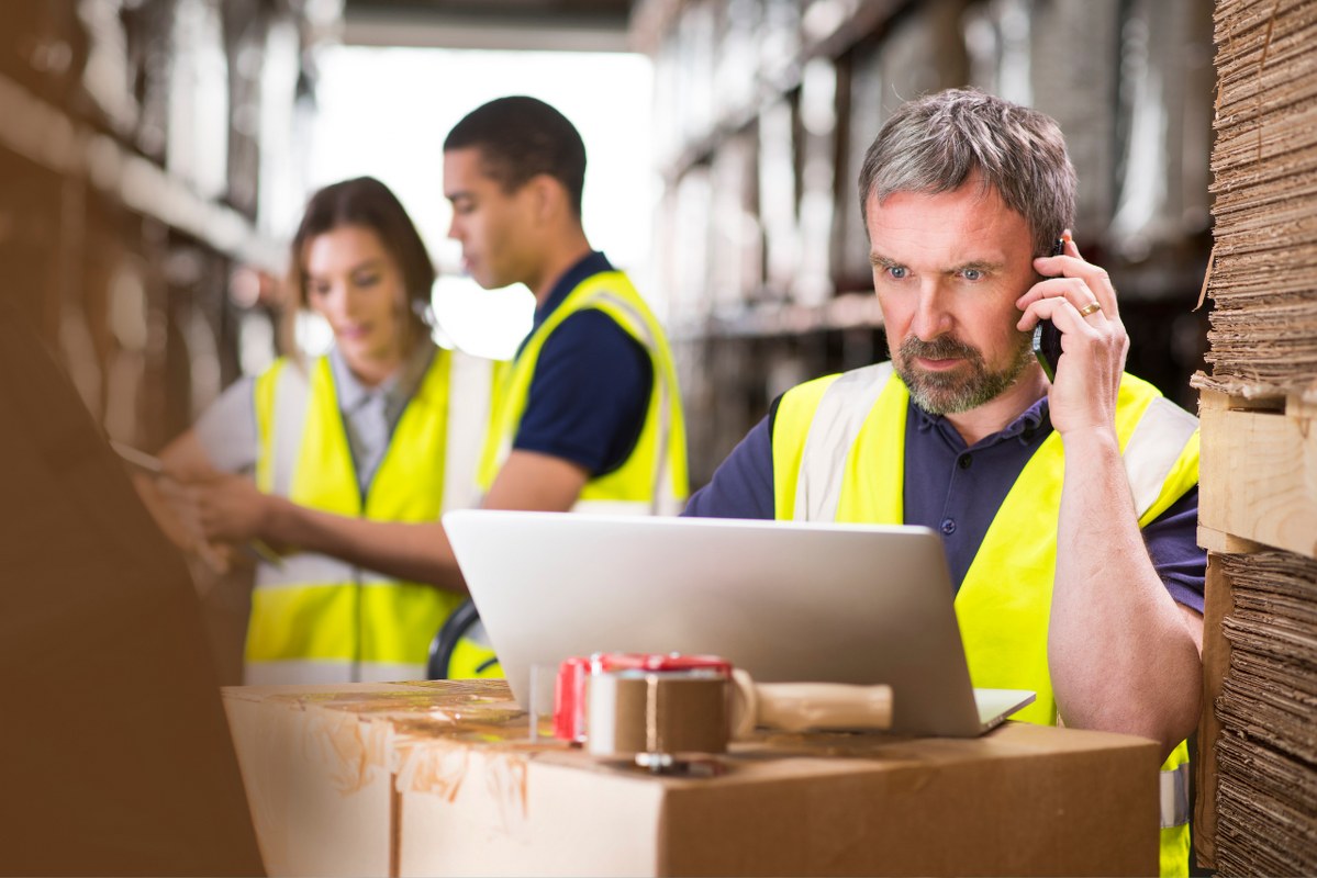Workers at the background while supplier is on the phone and laptop, inside warehouse while