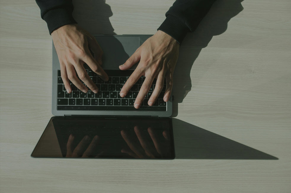 Hands typing on a laptop keyboard with a shadow cast across a light wooden desk.