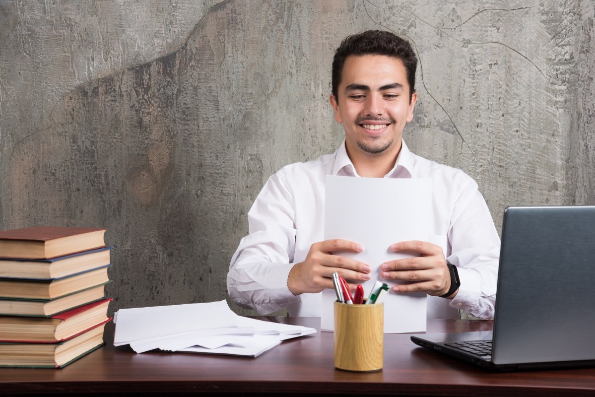 Man is holding some papers in front of his laptop.