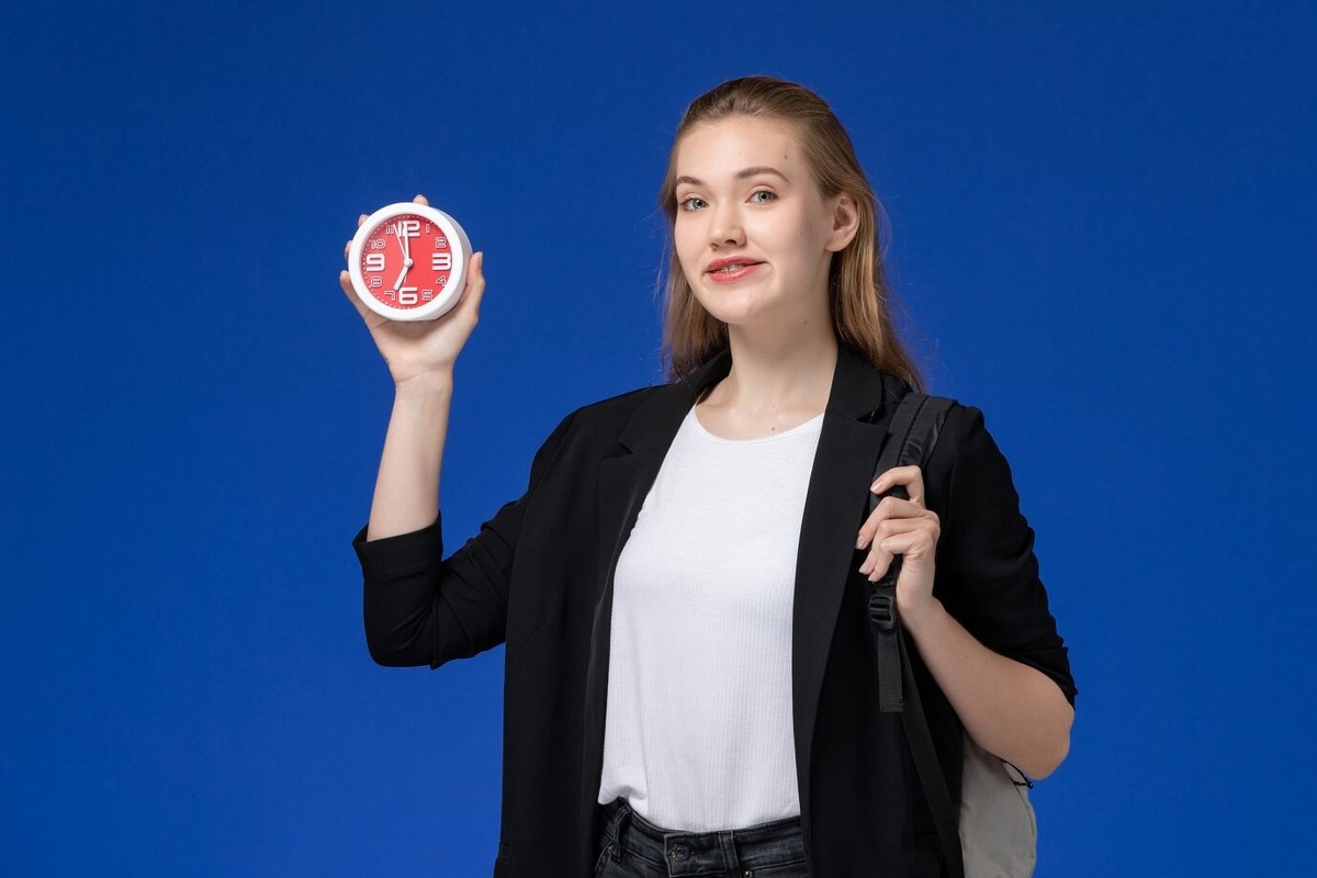 Student in black jacket wearing backpack holding a clock.