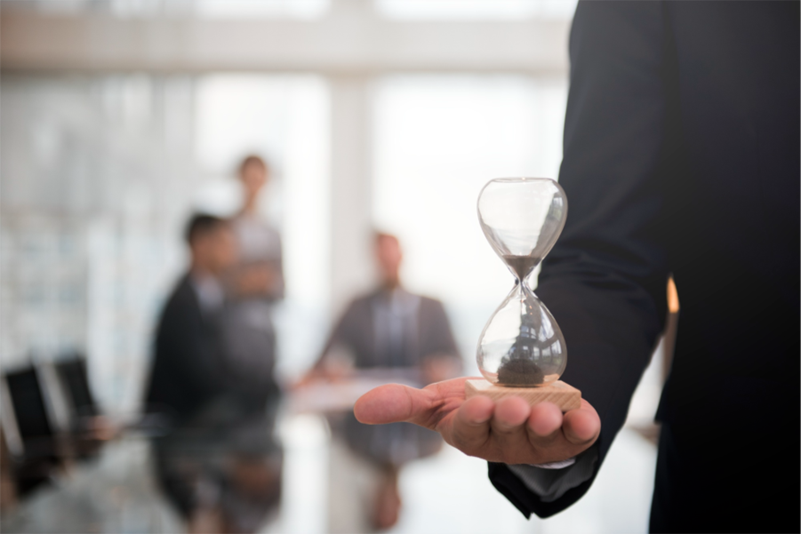 Businessperson holding an hourglass in a meeting room with blurred colleagues in the background, symbolizing time management and efficiency.