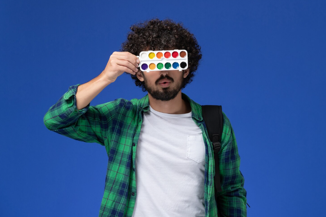 Front view of male student in green checkered shirt with black backpack holding paints for drawing.