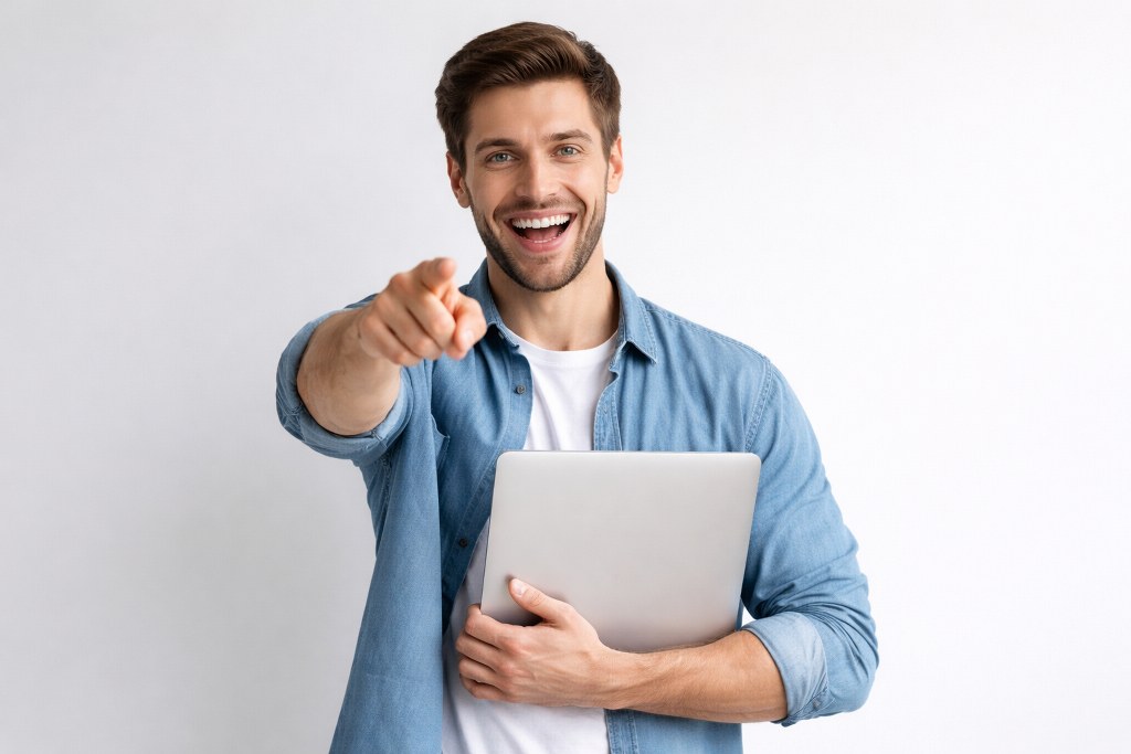 A smiling man points at the viewer while holding a laptop against his chest.